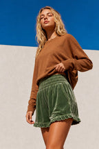 A woman wearing a brown long-sleeve knit sweater and green velvet shorts with a ruffled hem, standing against a split blue and white background.