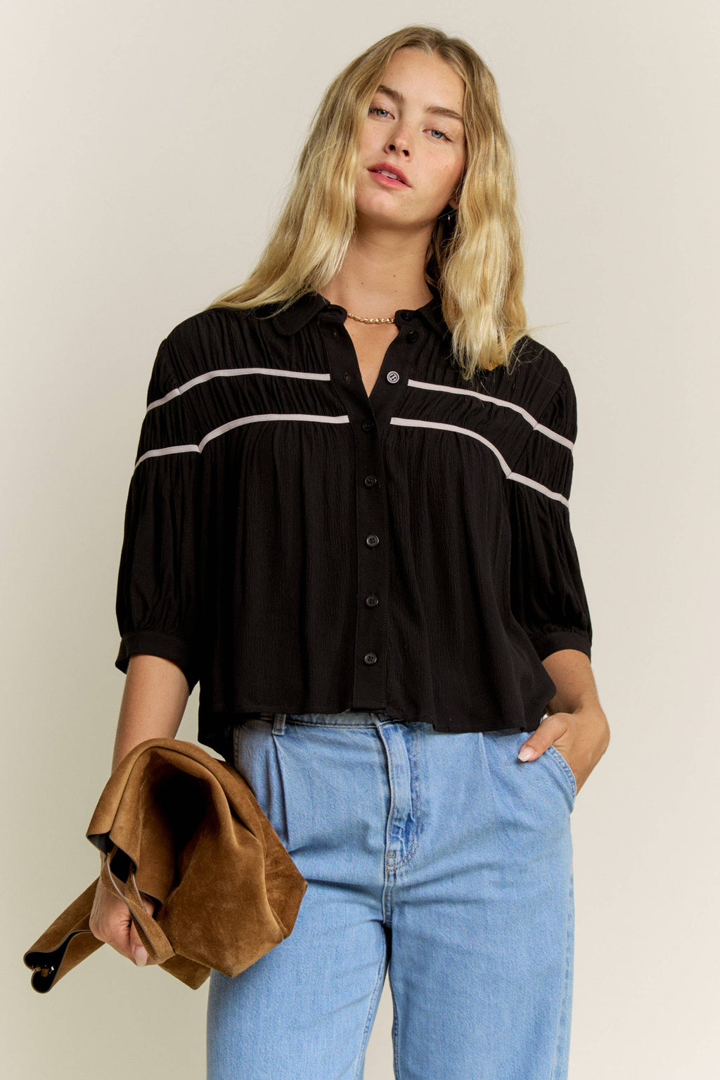 A woman modeling a black button-front blouse with horizontal white stripe details across the chest and shoulders, paired with light blue jeans and holding a brown suede clutch against a plain light beige studio background.