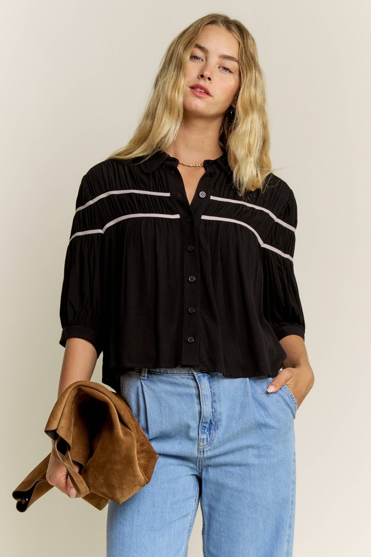 A woman modeling a black button-front blouse with horizontal white stripe details across the chest and shoulders, paired with light blue jeans and holding a brown suede clutch against a plain light beige studio background.