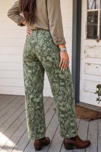 A person wearing green paisley wide-leg pants, a taupe long-sleeve shirt, and brown leather boots, standing on a wooden porch in front of a white door.