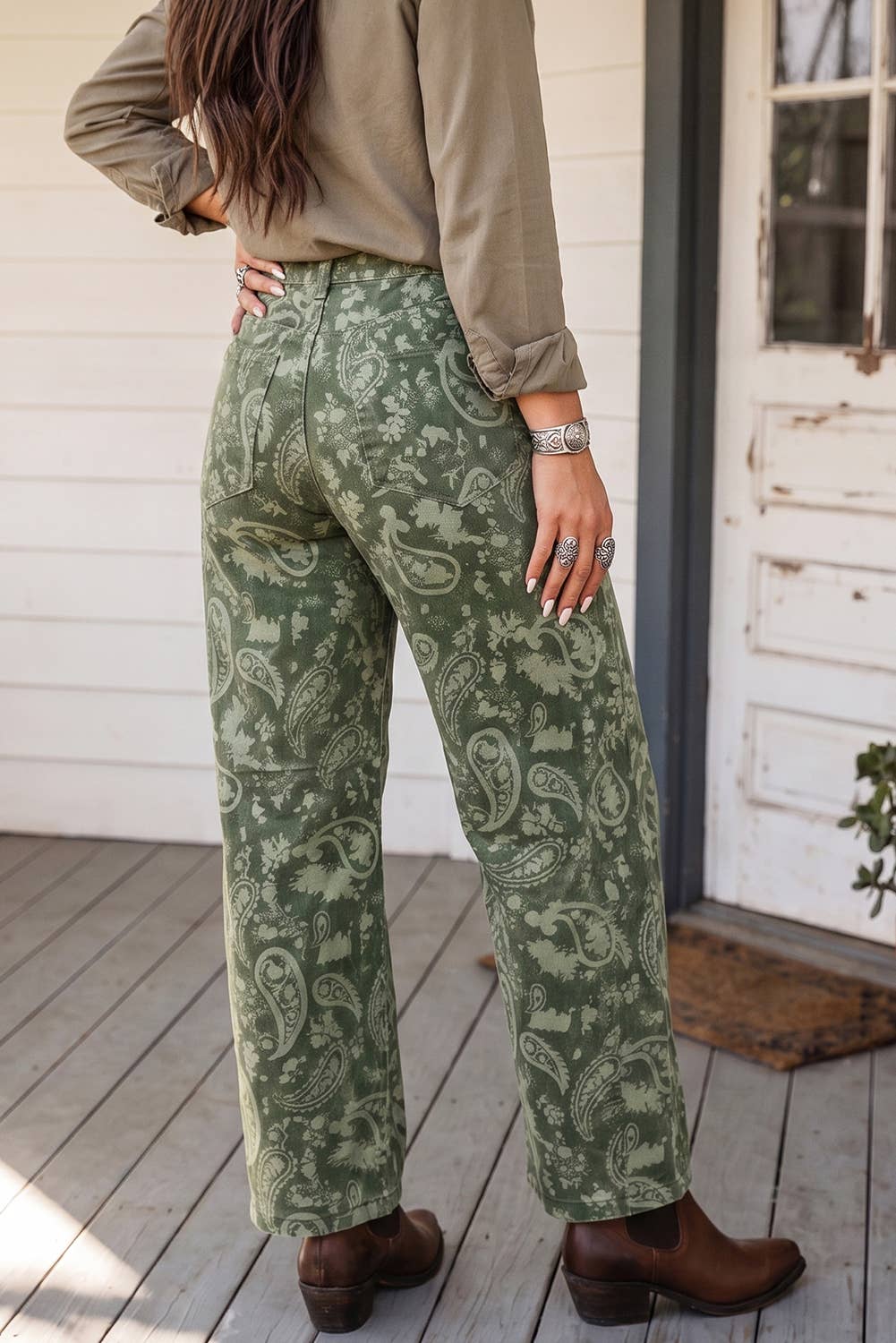A person wearing green paisley wide-leg pants, a taupe long-sleeve shirt, and brown leather boots, standing on a wooden porch in front of a white door.