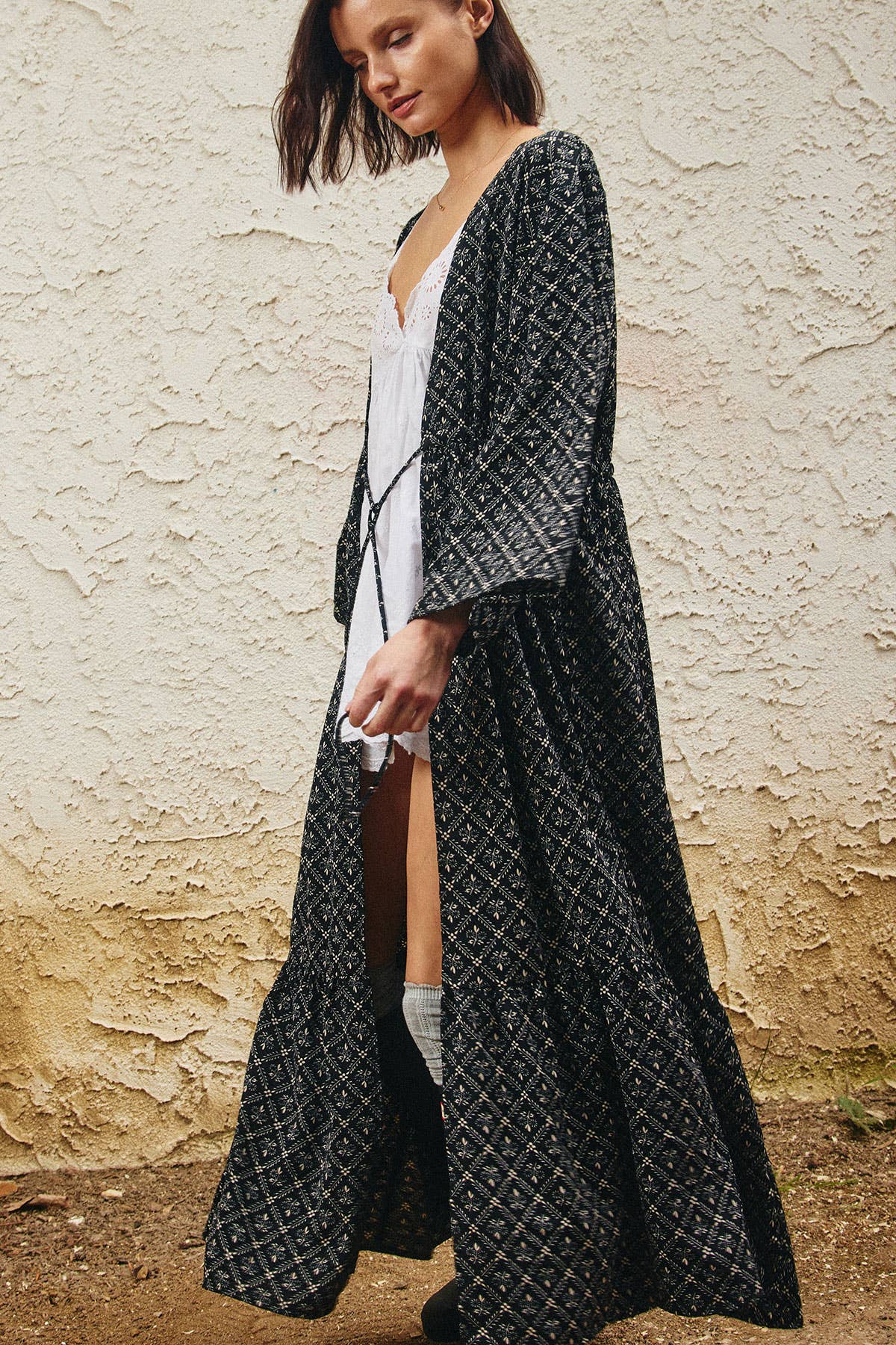 A woman wearing a long black kimono-style robe with a white geometric pattern over a white dress, standing against a textured beige stucco wall.