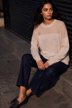 A woman sits on a concrete curb against a dark metal shutter, wearing a cream-colored knit sweater with a solid front panel and sheer mesh sleeves, paired with dark blue jeans and black loafers.
