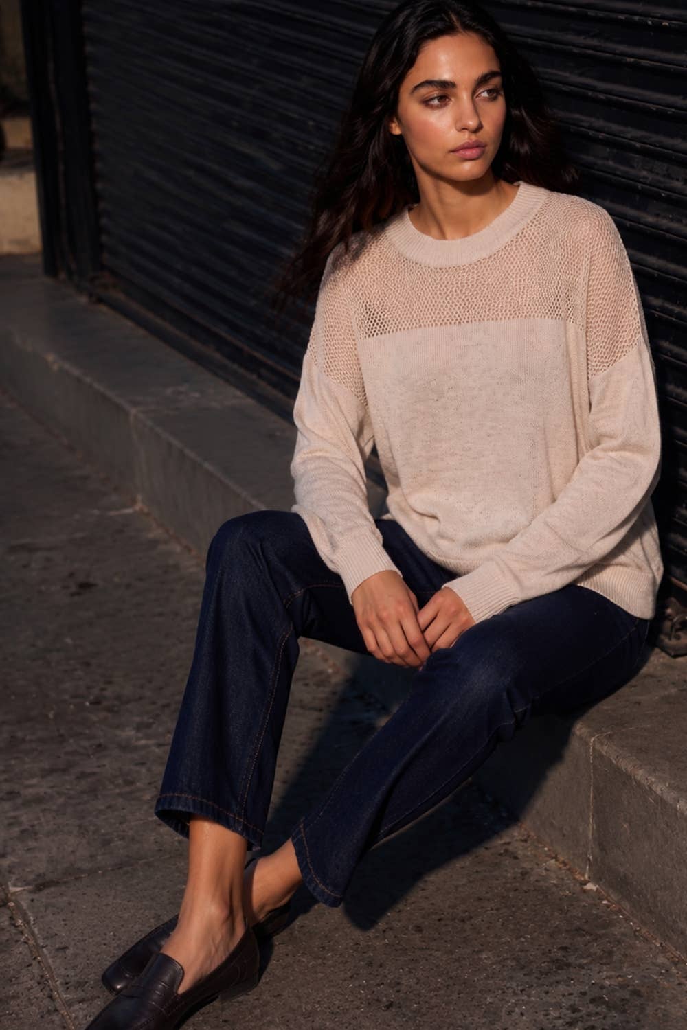A woman sits on a concrete curb against a dark metal shutter, wearing a cream-colored knit sweater with a solid front panel and sheer mesh sleeves, paired with dark blue jeans and black loafers.