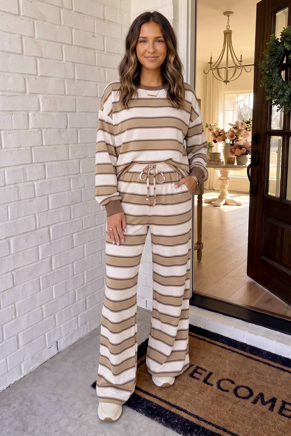 A woman wearing a matching beige and cream striped lounge set with a long-sleeve top and wide-leg pants, standing on a porch in front of a white brick wall.