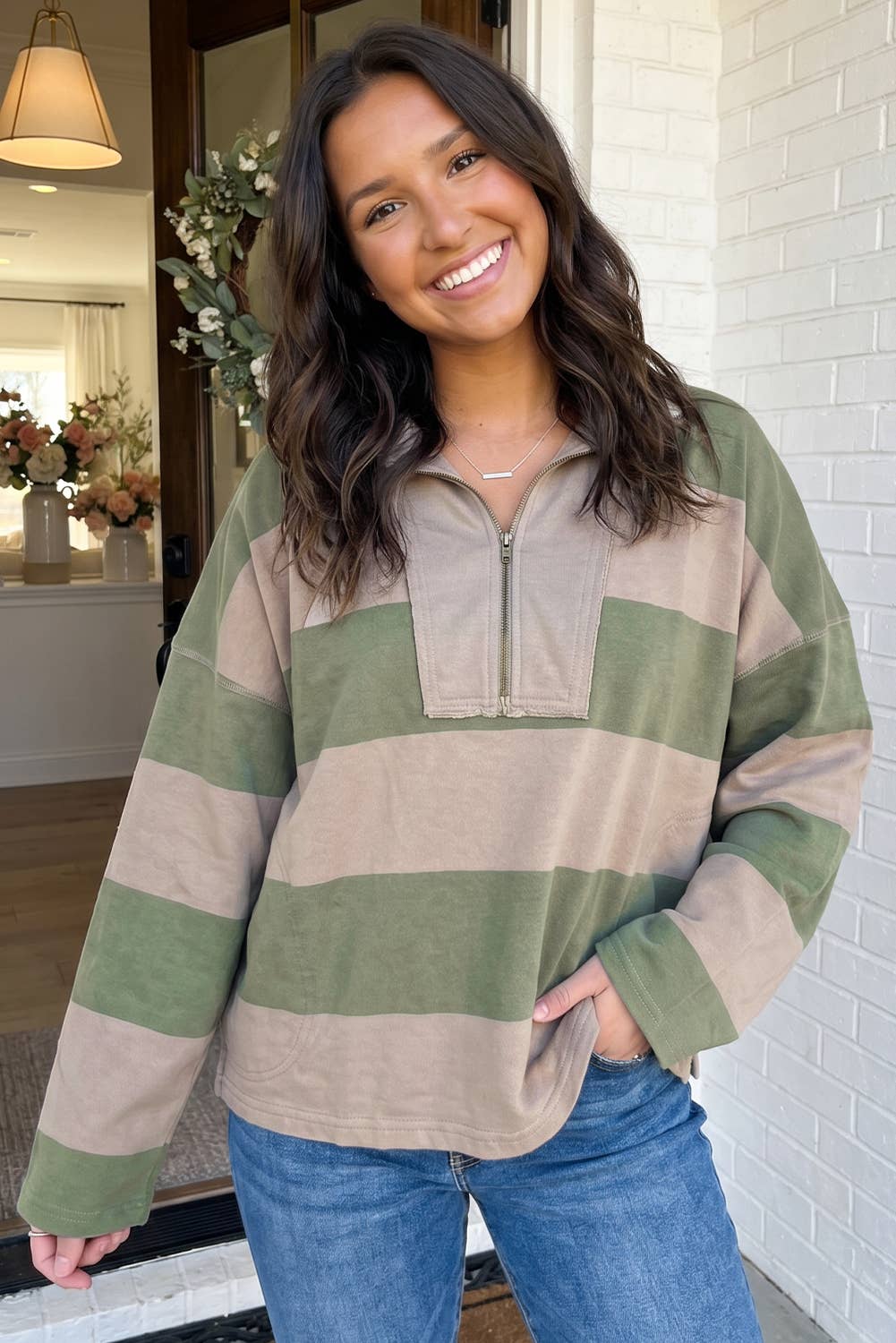 A woman smiling while wearing a long-sleeve quarter-zip pullover sweater with wide horizontal stripes in sage green and beige, paired with blue jeans.