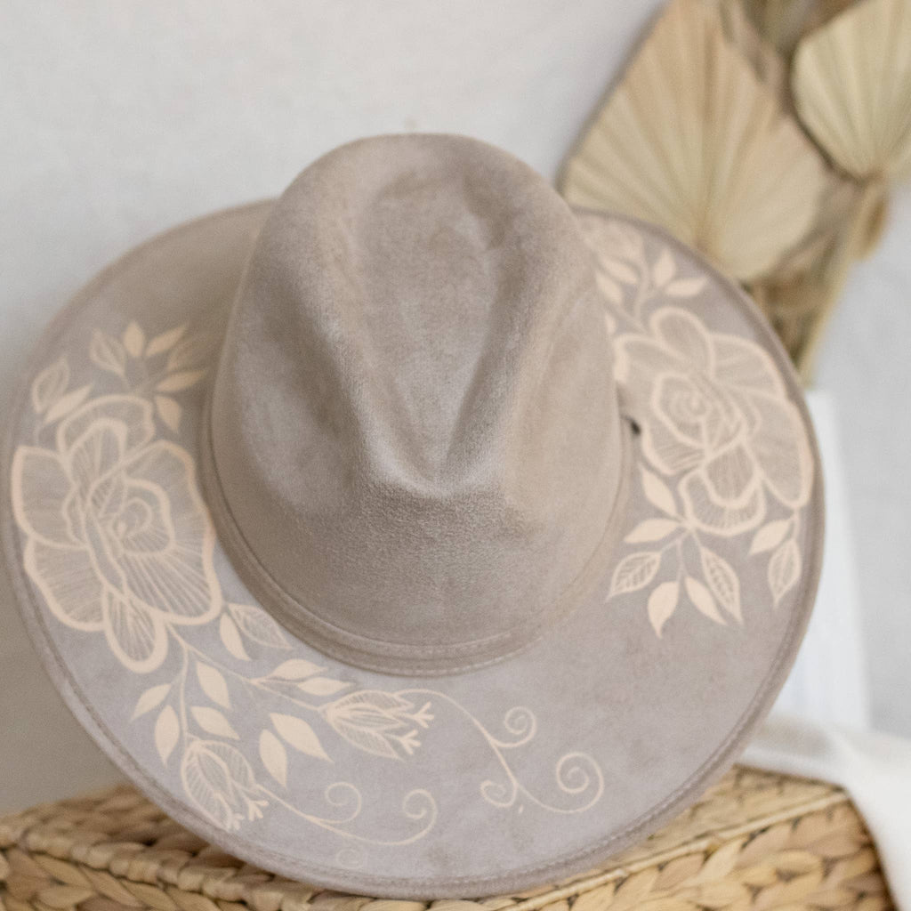 A taupe wide-brim fedora hat with artisan painted beige floral print on the brim, displayed on a woven straw mat. Crafted in Oaxaca, Mexico.