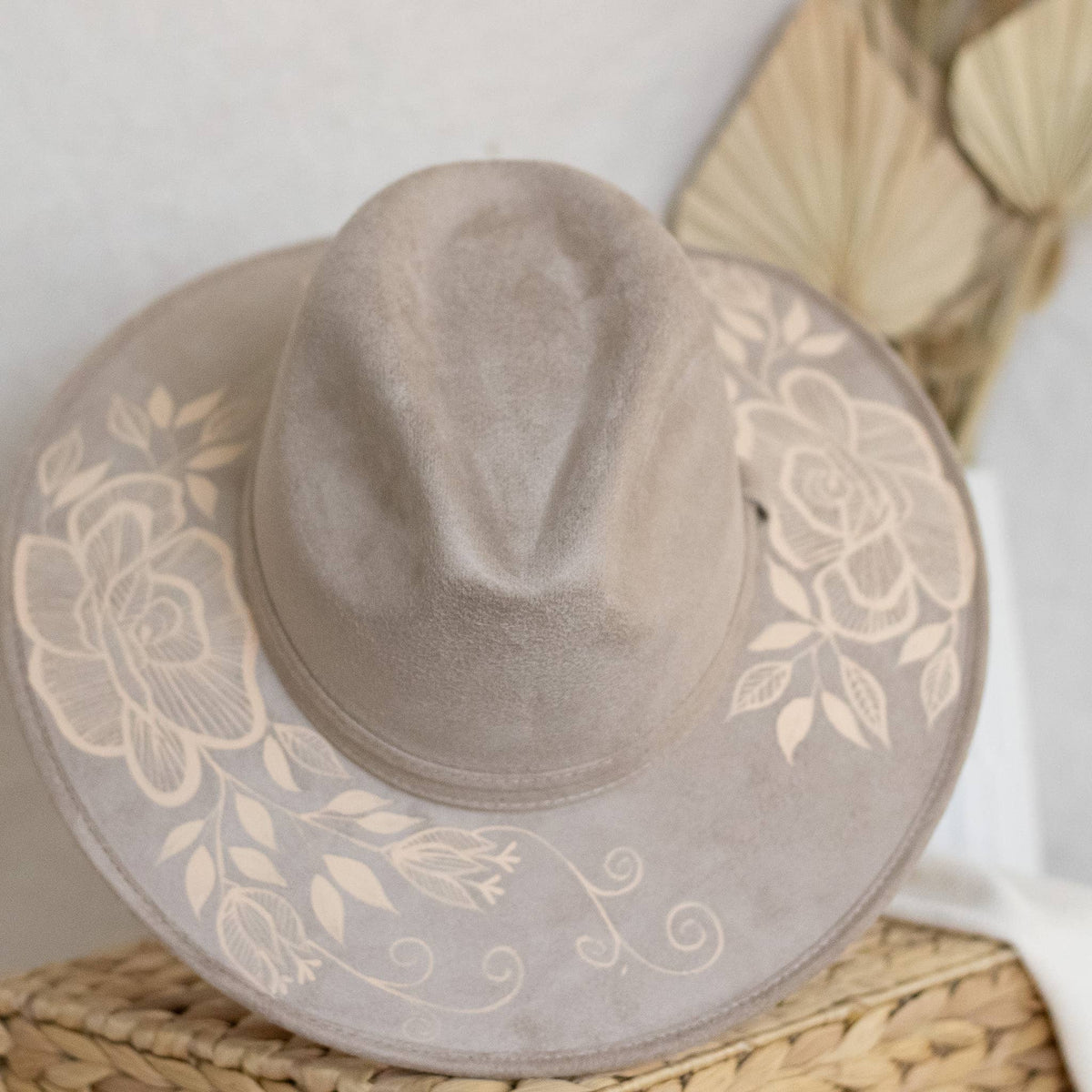 A taupe wide-brim fedora hat with artisan painted beige floral print on the brim, displayed on a woven straw mat. Crafted in Oaxaca, Mexico.