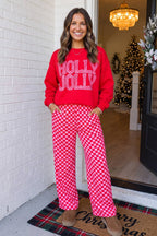 A woman stands on a front porch wearing a bright red crewneck sweatshirt with 'HOLLY JOLLY' in white glitter-style lettering, paired with pink and white checkered wide-leg pants and brown suede boots.