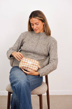Seated woman in a gray sweater holding the terracotta and mustard gingham squared makeup bag on her lap