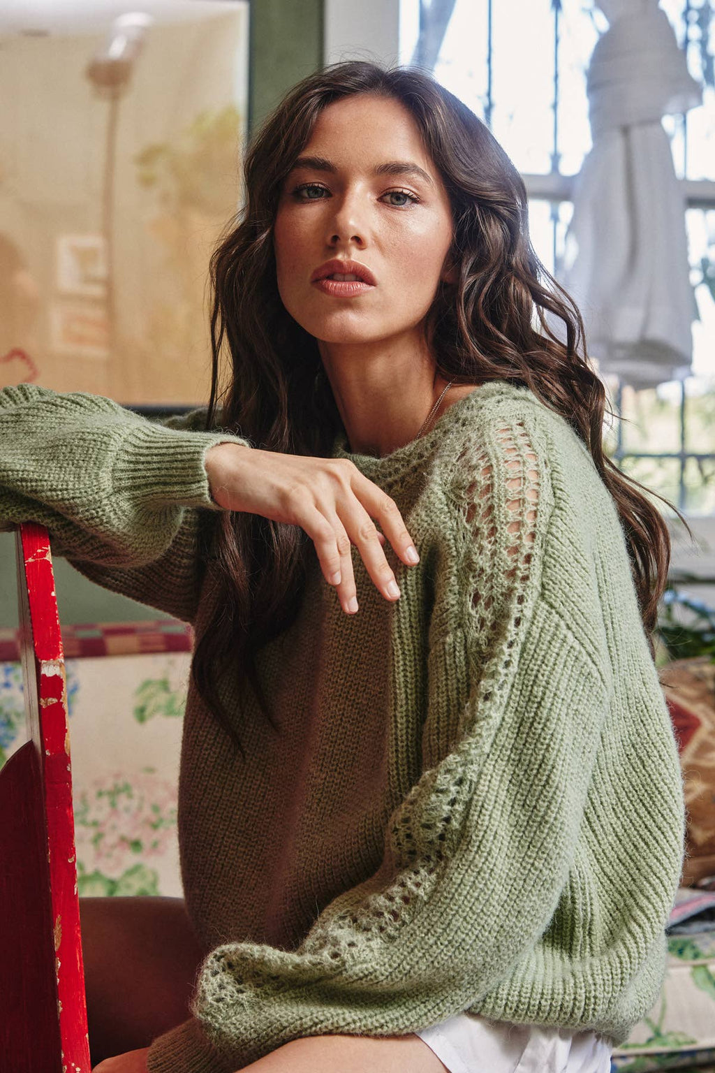 Woman wearing a moss green knit pullover sweater with crochet details on the boat neckline and down the sleeves