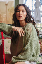 Woman wearing a moss green knit pullover sweater with crochet details on the boat neckline and down the sleeves