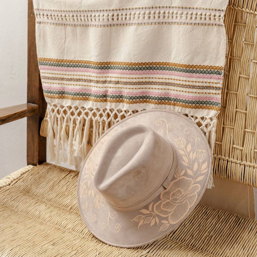 A light beige wide-brim fedora hat with artisan painted floral print rests on a woven rattan chair, with a matching cream and multicolor woven textile draped over the chair. Crafted in Oaxaca, Mexico.
