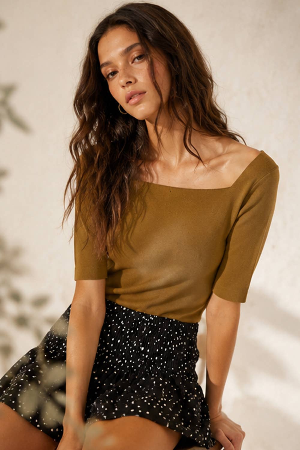 A woman wearing a fitted short-sleeve top in a warm olive-brown color paired with a black skirt featuring small white polka dots, posed against a softly lit neutral wall.