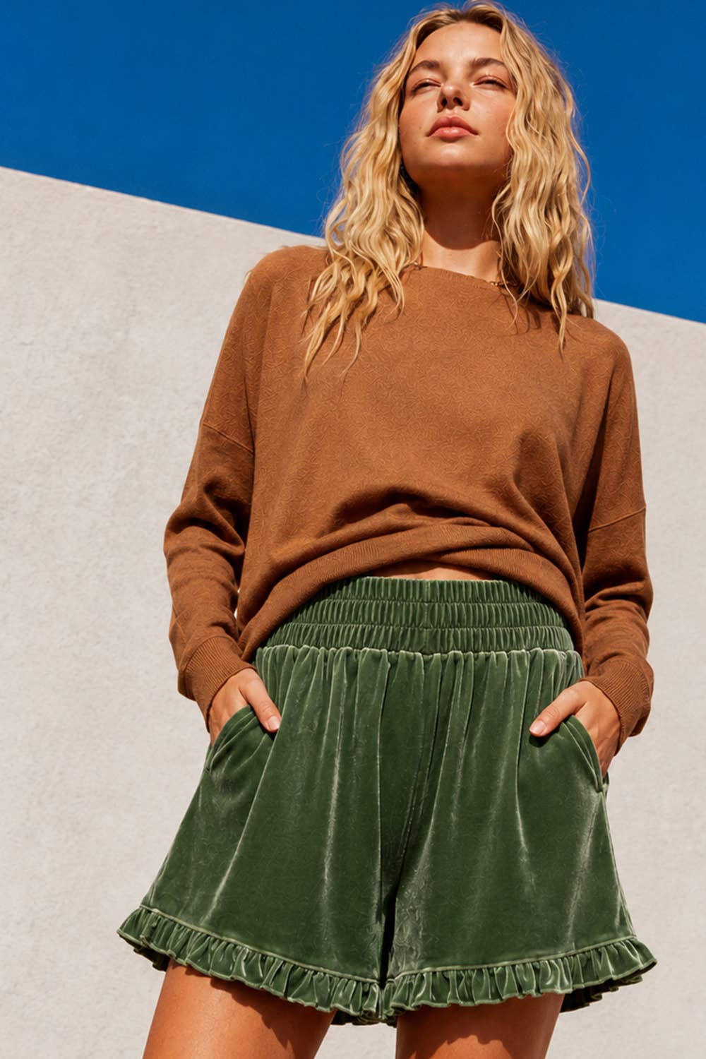A woman wearing a brown knit sweater and green velvet shorts, standing outdoors against a split gray and blue background.