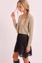 A woman modeling a beige long-sleeve knit cardigan with a tie-front detail, paired with a black skirt featuring a lace hem, against a solid pale pink background.