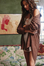 A woman wearing a brown fuzzy knit cardigan over a white camisole and shorts, standing in a room with a floral-patterned sofa and a green wall.