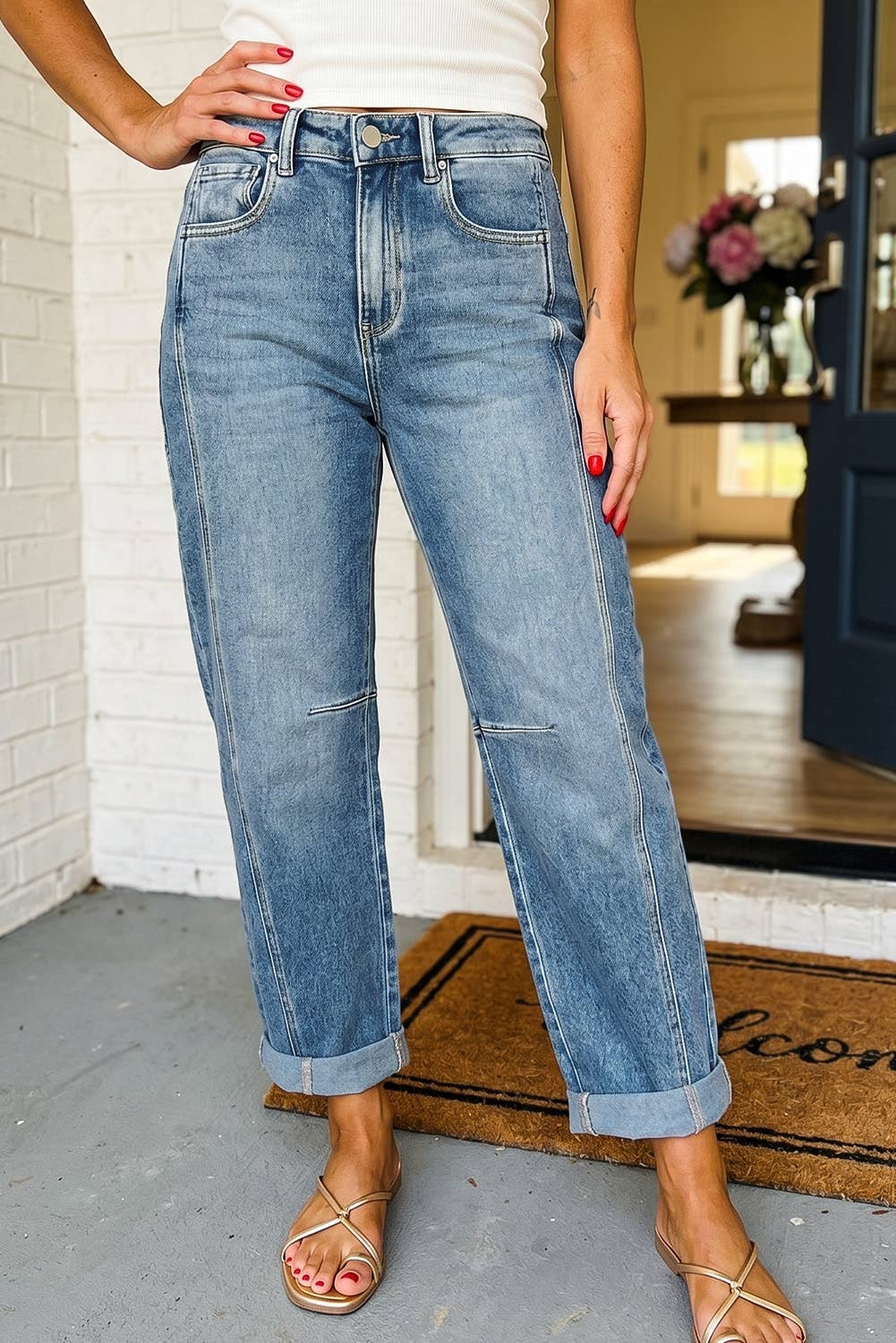 A person wearing light-wash blue cropped jeans with rolled cuffs, paired with a white top and strappy flat sandals, standing on a porch in front of a white brick wall.