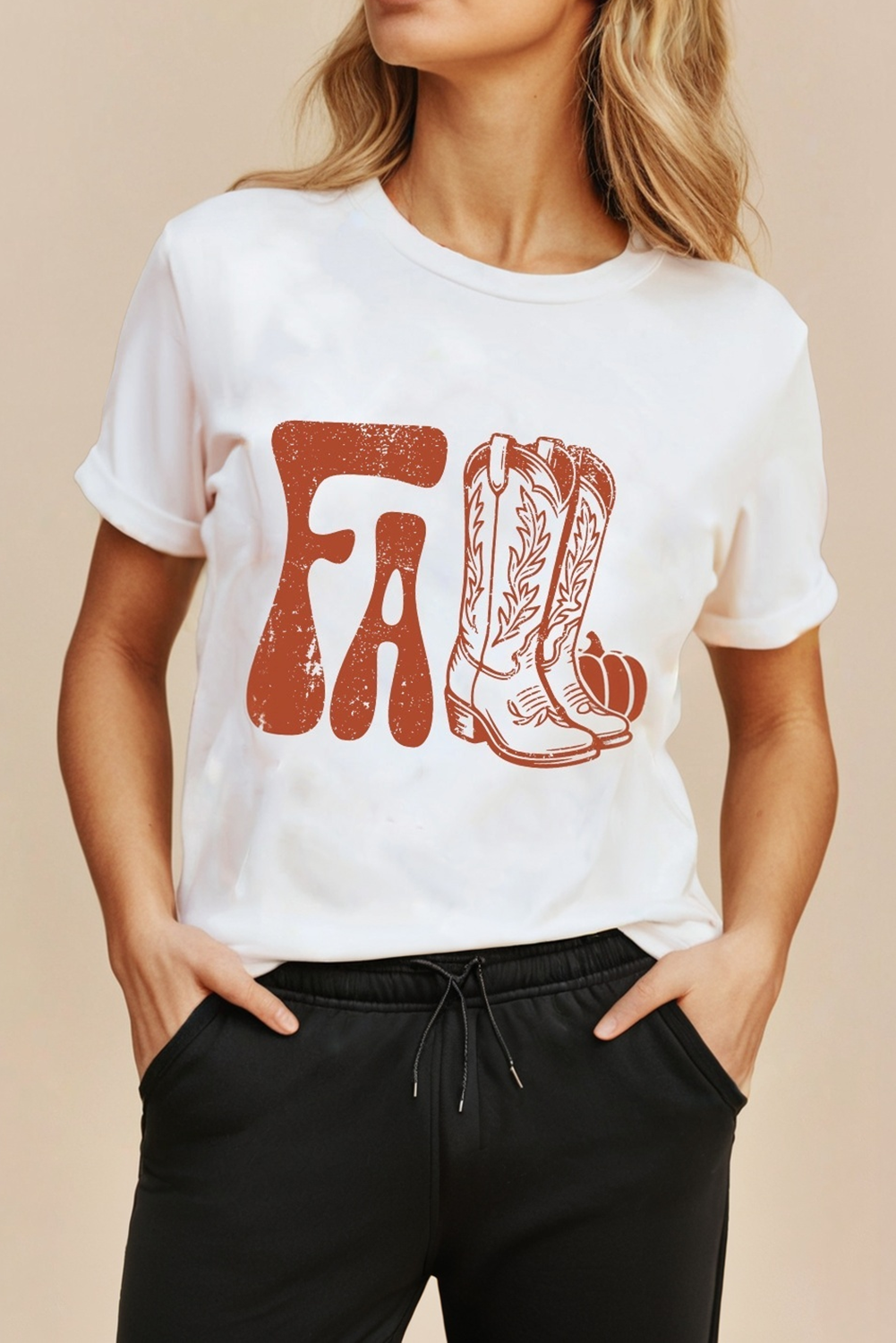 A woman wearing a white short-sleeve crewneck t-shirt with a distressed orange graphic spelling 'FA' in the shape of cowboy boots and pumpkins, paired with black sweatpants against a solid beige background.