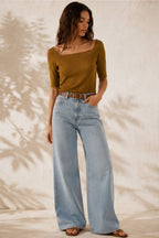 A woman modeling a short-sleeve olive-brown top and light-wash wide-leg jeans with a brown belt, standing against a textured beige wall with leaf shadows.