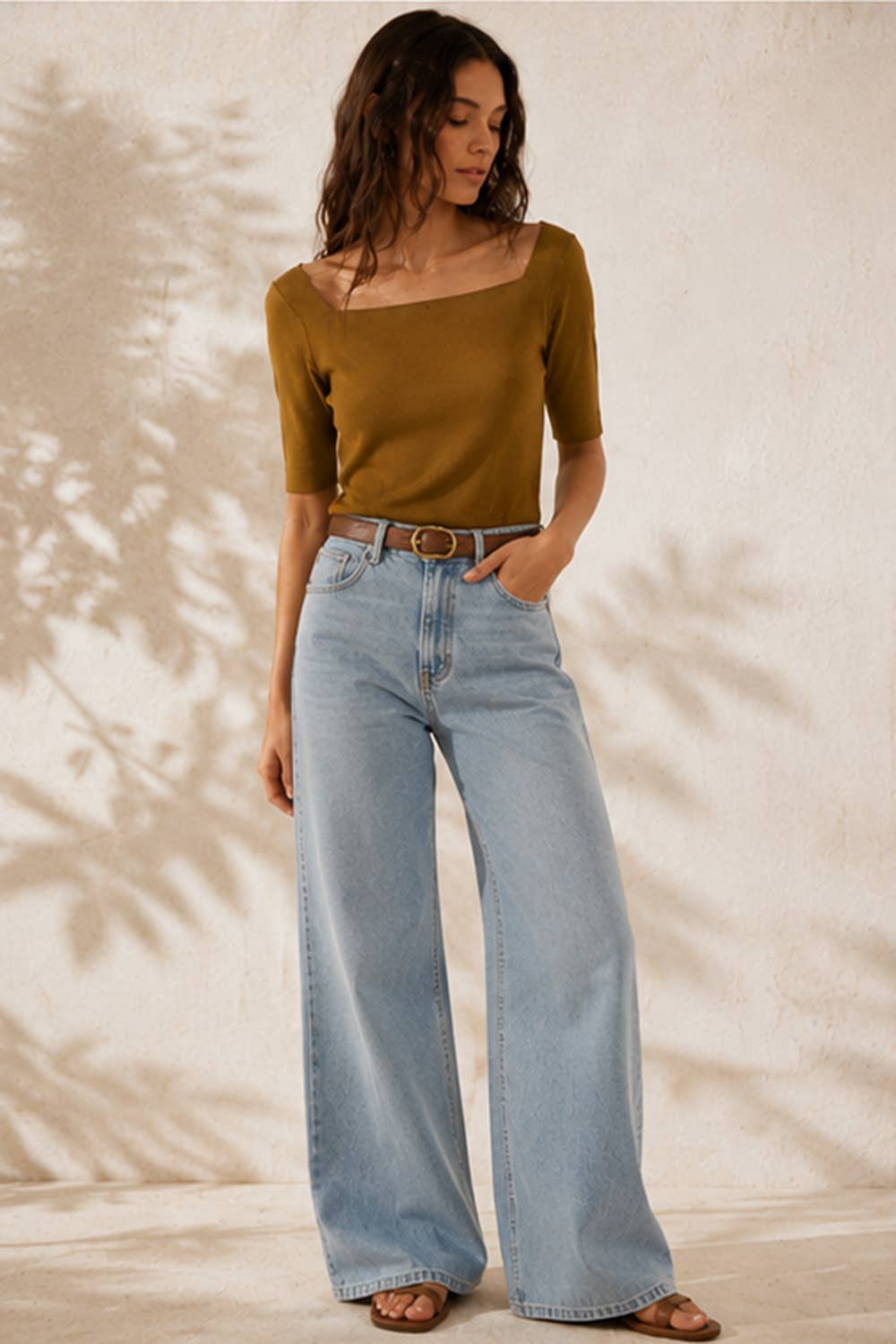 A woman modeling a short-sleeve olive-brown top and light-wash wide-leg jeans with a brown belt, standing against a textured beige wall with leaf shadows.