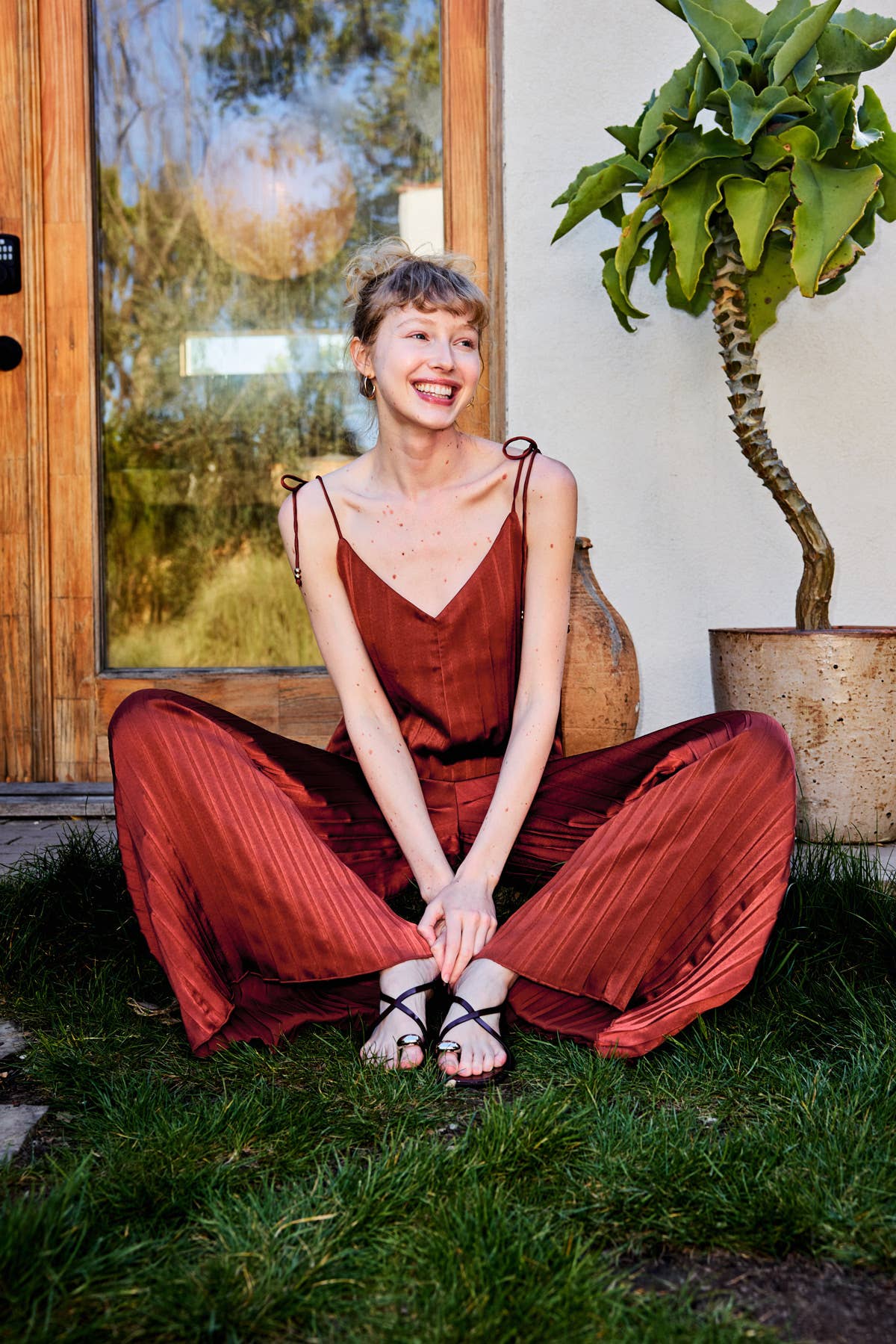 A woman sitting cross-legged on green grass in front of a wooden door and potted plant, wearing a rust-brown pleated camisole and matching wide-leg pants.