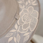 Close-up of a taupe suede hat featuring a large artisan painted cream floral and leaf pattern printed or embroidered on the side. Crafted in Oaxaca, Mexico.