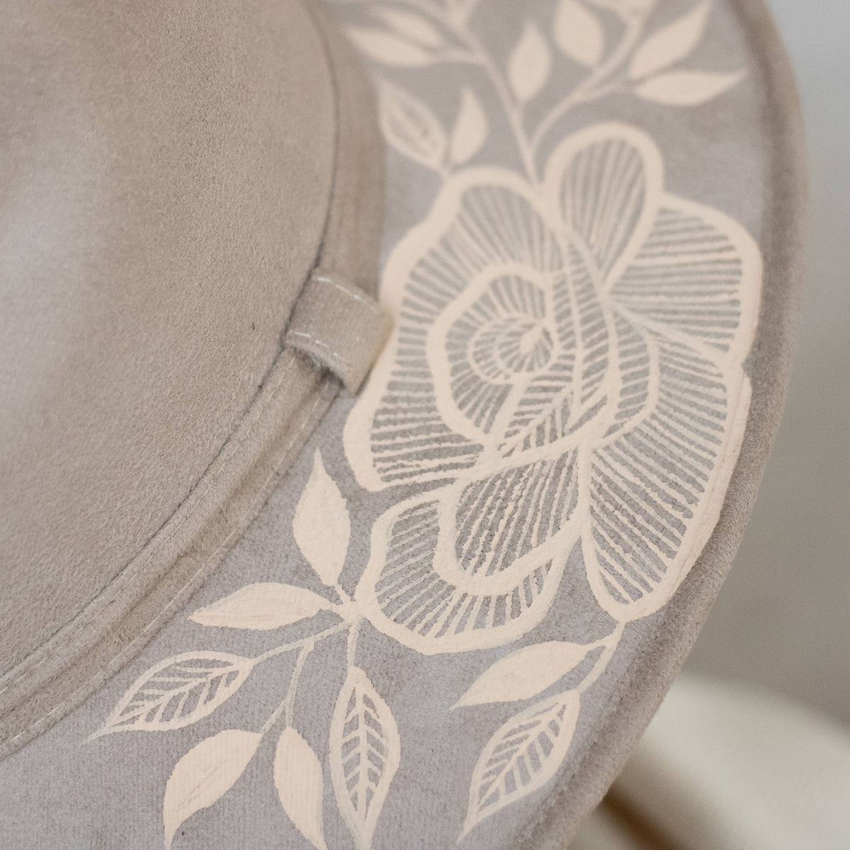 Close-up of a taupe suede hat featuring a large artisan painted cream floral and leaf pattern printed or embroidered on the side. Crafted in Oaxaca, Mexico.