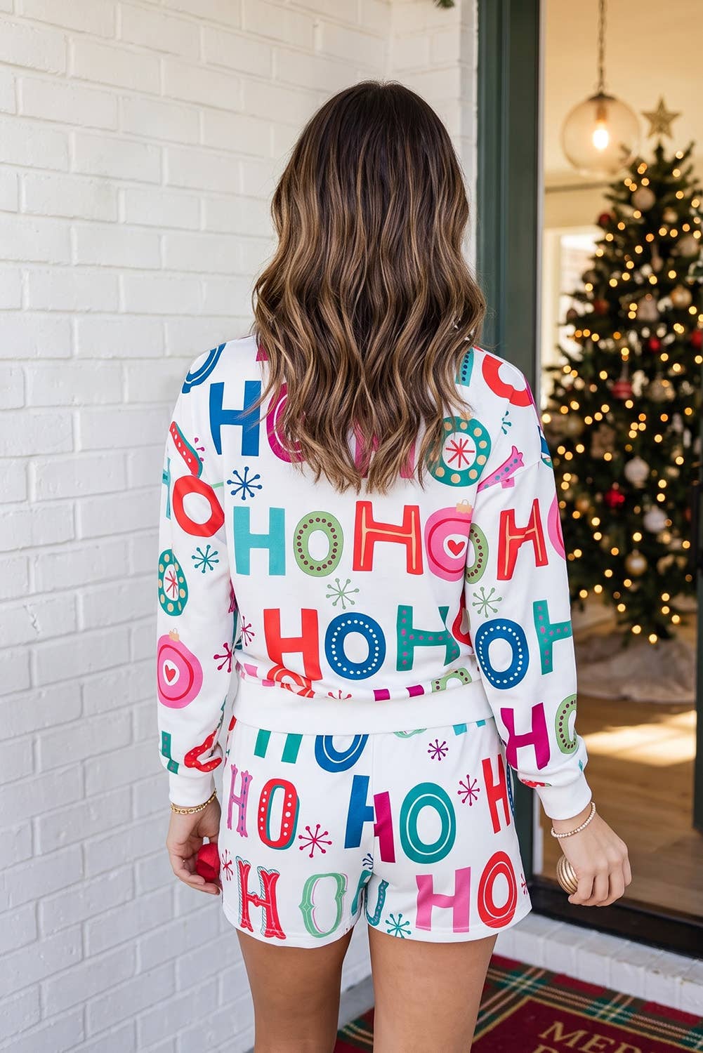 A woman viewed from behind wearing a matching white Christmas-themed sweatshirt and shorts set with a colorful 'HO HO HO' letter print, standing in front of a white brick wall and a decorated Christmas tree.