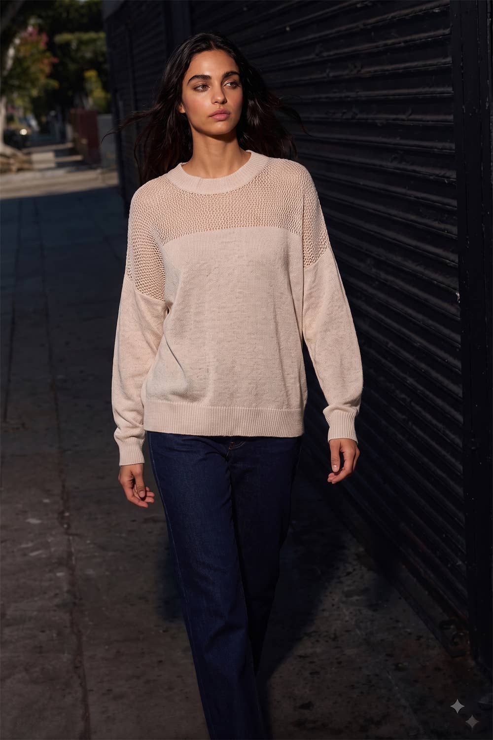 A woman wearing a light beige knit crewneck sweater with textured open-knit sleeves and a solid knit body, paired with dark blue jeans, walking outdoors against a dark corrugated metal wall.