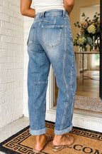A person wearing light blue wide-leg cropped jeans with rolled cuffs and back pocket details, standing indoors near a white brick wall and doorway.