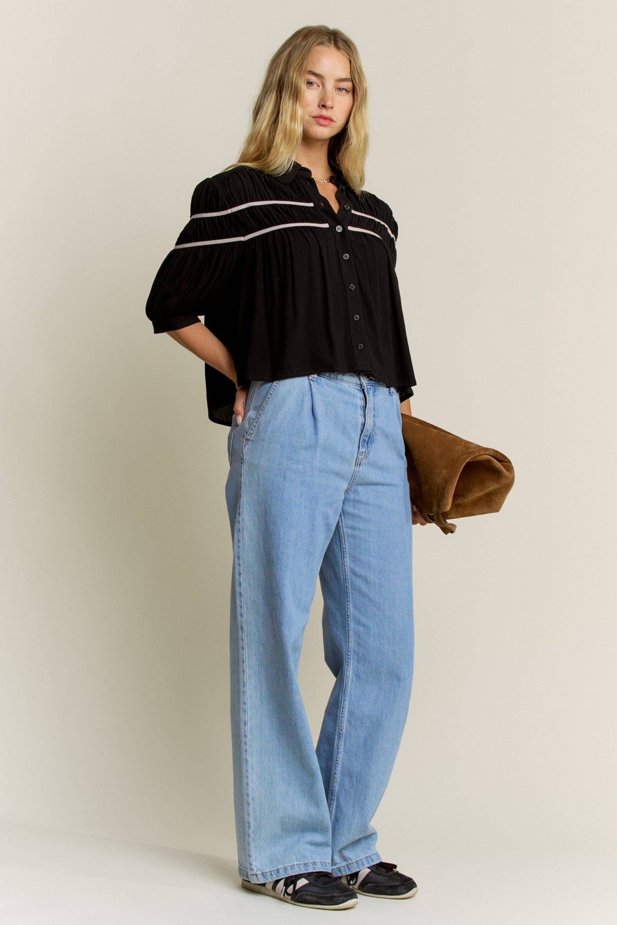 A woman modeling a black short-sleeve button-up blouse with white stripe details on the shoulders, paired with light blue wide-leg jeans and holding a brown clutch bag against a plain beige studio background.