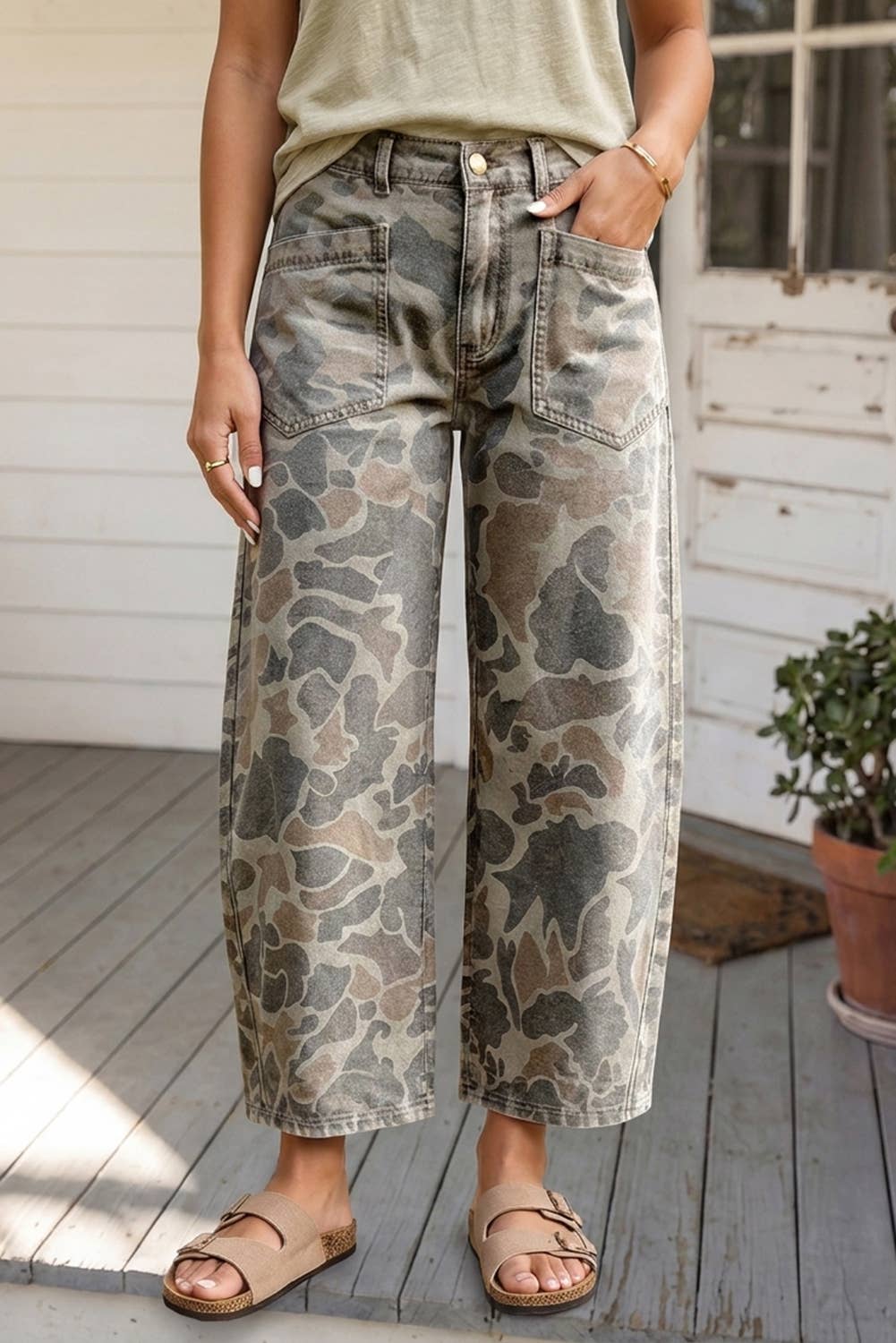 A person wearing wide-leg cropped jeans with a muted camouflage pattern in beige, gray, and brown, paired with a light olive top and tan slide sandals, standing on a weathered wooden porch.