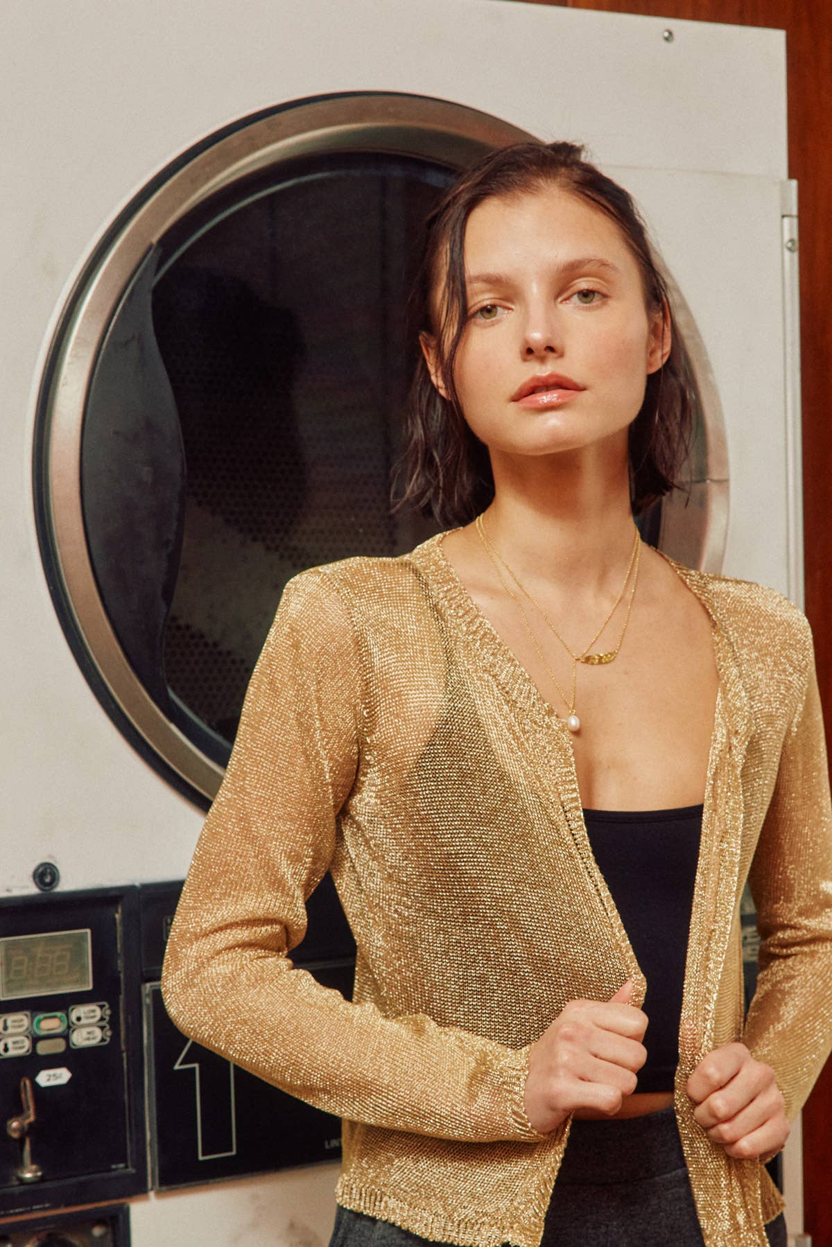 A woman models a shimmering gold textured cardigan over a black top, standing in front of a large washing machine.