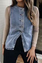 A woman wearing a sleeveless light-wash denim vest over a beige long-sleeve top and black pants, standing indoors against a light brick wall.