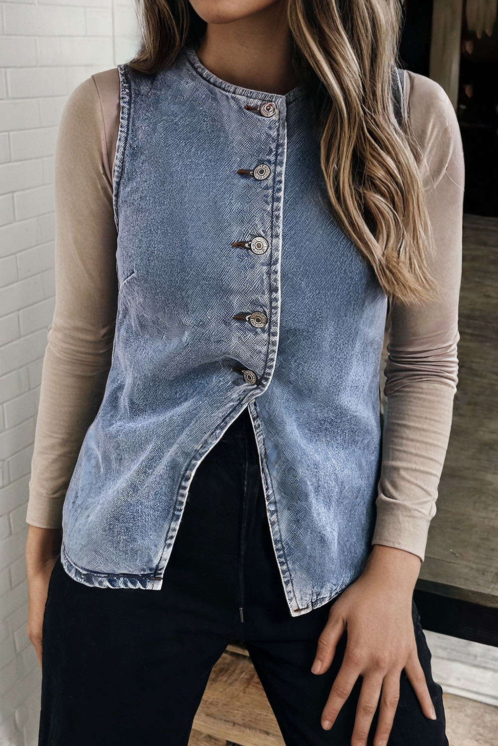 A woman wearing a sleeveless light-wash denim vest over a beige long-sleeve top and black pants, standing indoors against a light brick wall.