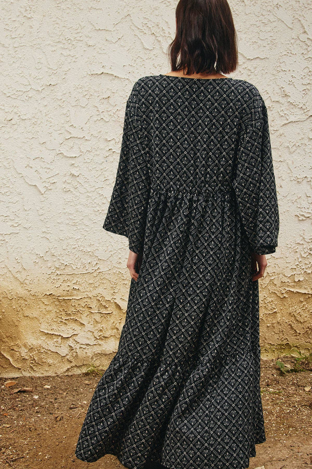 A woman viewed from behind wearing a long-sleeved black maxi dress with a white geometric pattern, standing in front of a textured beige stucco wall.