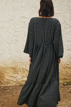 A woman viewed from behind wearing a long-sleeved black maxi dress with a white geometric pattern, standing in front of a textured beige stucco wall.