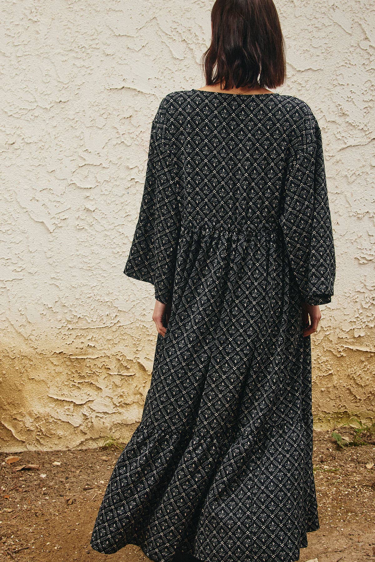 A woman viewed from behind wearing a long-sleeved black maxi dress with a white geometric pattern, standing in front of a textured beige stucco wall.