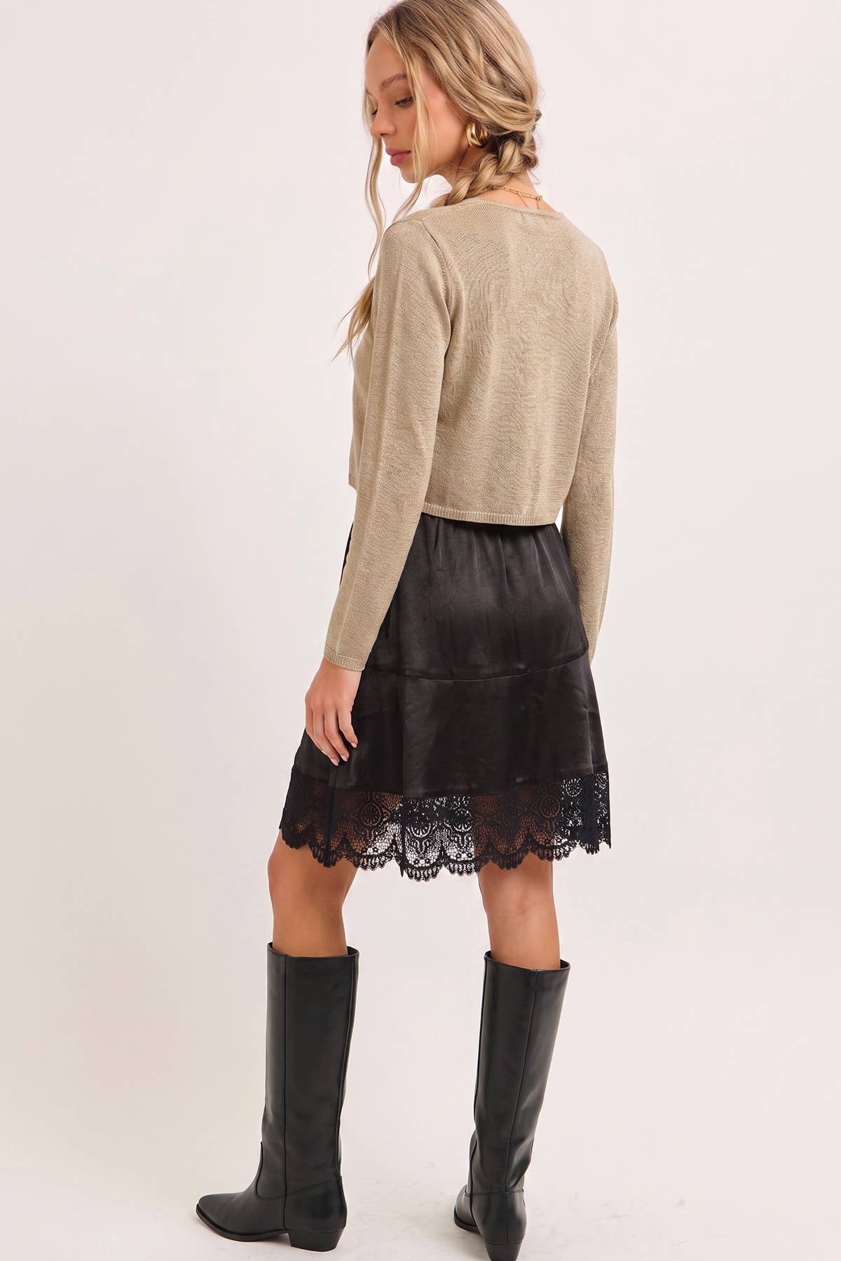A woman shown from the back wearing a beige long-sleeve cropped sweater, a black satin A-line skirt with a lace hem, and black knee-high boots, against a plain light background.