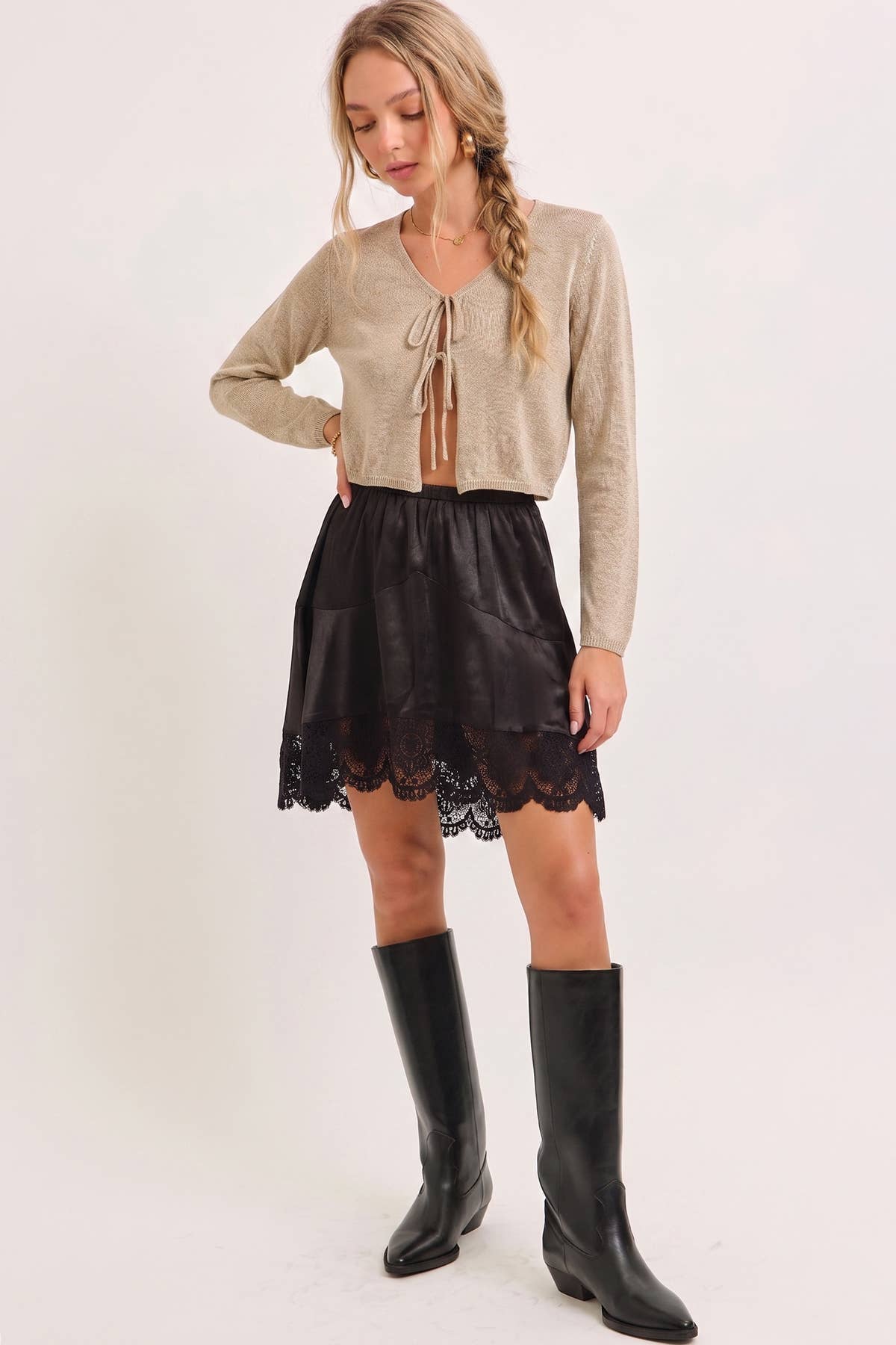 A woman modeling a beige knit long-sleeve crop top with a tie neckline, paired with a black skirt featuring a lace hem and black knee-high boots, against a plain white studio background.