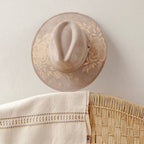 A beige wide-brim fedora hat with artisan painted embroidered floral patterns on the brim, hanging on a textured off-white wall above a woven rattan chair and a cream embroidered tablecloth. Crafted in Oaxaca, Mexico.