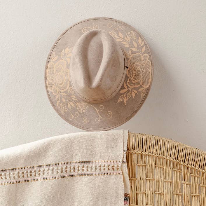 A beige wide-brim fedora hat with artisan painted embroidered floral patterns on the brim, hanging on a textured off-white wall above a woven rattan chair and a cream embroidered tablecloth. Crafted in Oaxaca, Mexico.