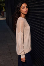 A woman wearing a light beige knit sweater with open-knit shoulder panels, standing outdoors against a dark corrugated metal wall.