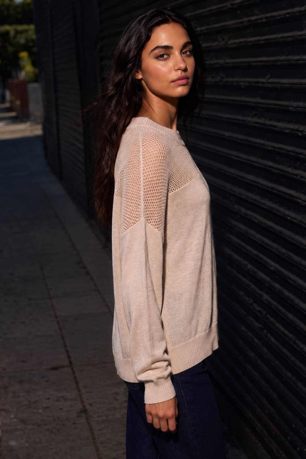 A woman wearing a light beige knit sweater with open-knit shoulder panels, standing outdoors against a dark corrugated metal wall.