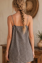 A woman shown from behind wearing a black and white gingham slip dress with thin straps and side ties, standing in a rustic indoor setting with a straw hat and wooden furniture in the background.