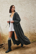 A woman wearing a long black patterned cardigan over a white lace dress, gray socks, and black rain boots, standing against a textured beige stucco wall.