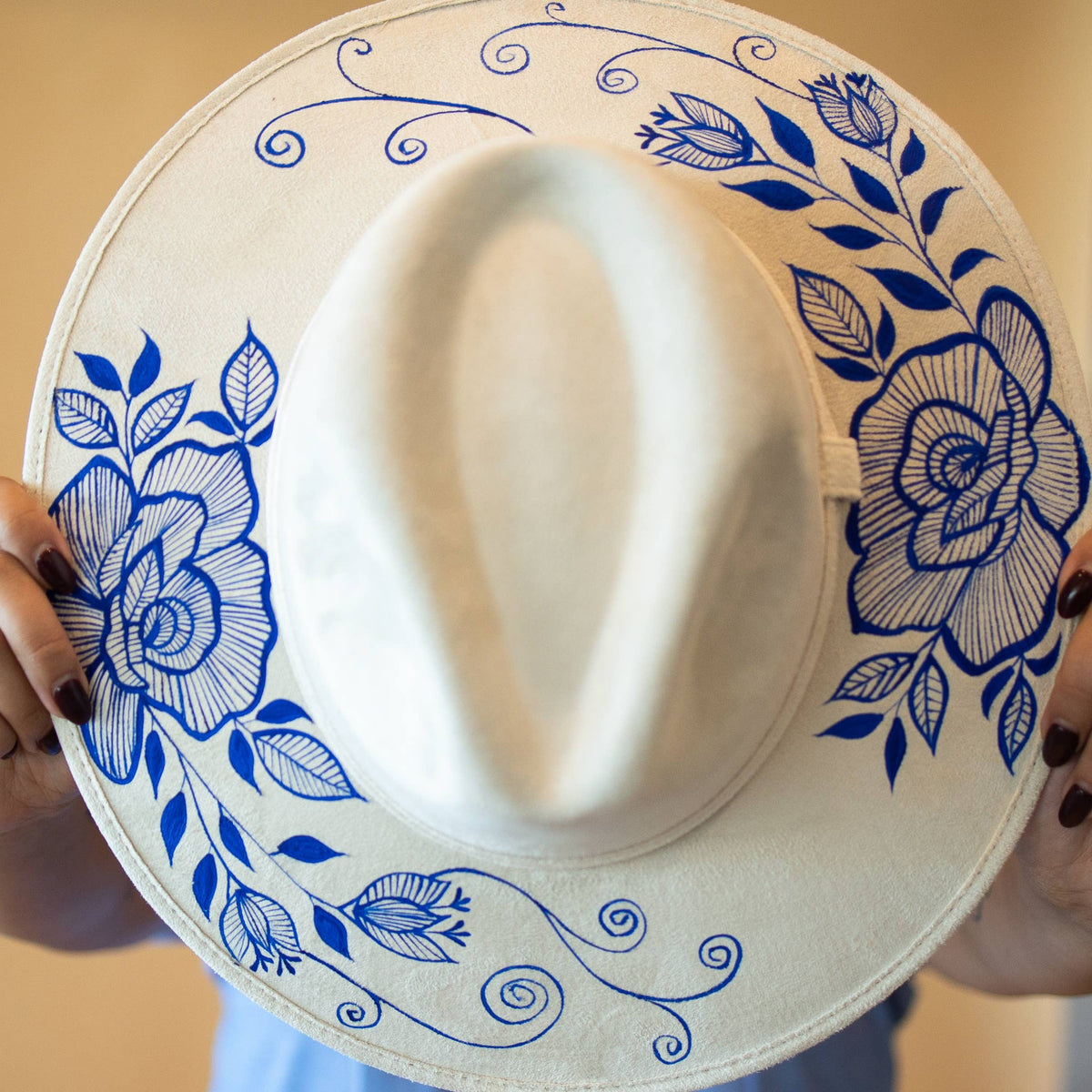 Made in Oaxaca, Mexico: a white hat with artisan painted blue floral patterns held by a person against a beige background