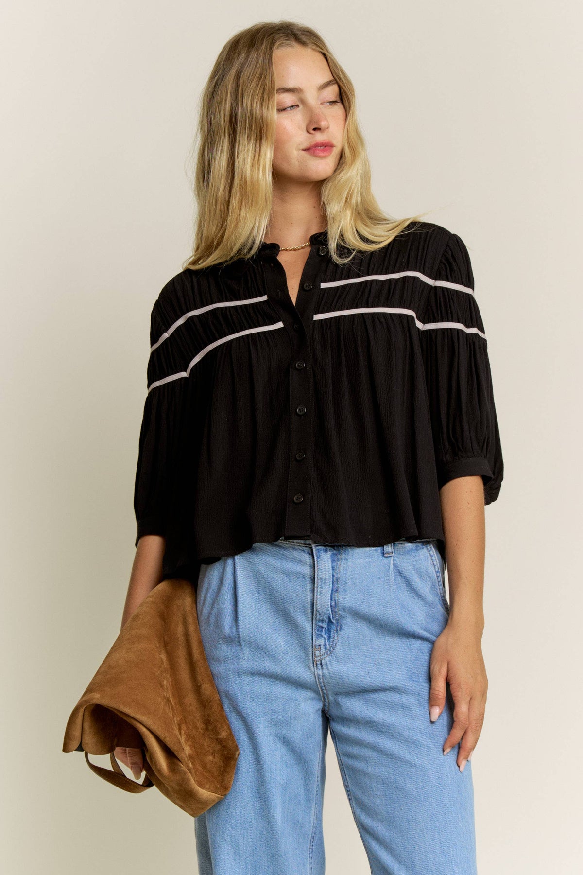 A woman wearing a black pleated blouse with white horizontal stripes on the sleeves, paired with light blue jeans and holding a brown suede clutch bag, set against a plain beige studio background.