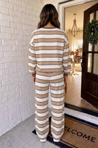 A woman viewed from behind wearing a matching beige and off-white striped lounge set, standing at a doorway with a brick wall on the left and a doormat visible.